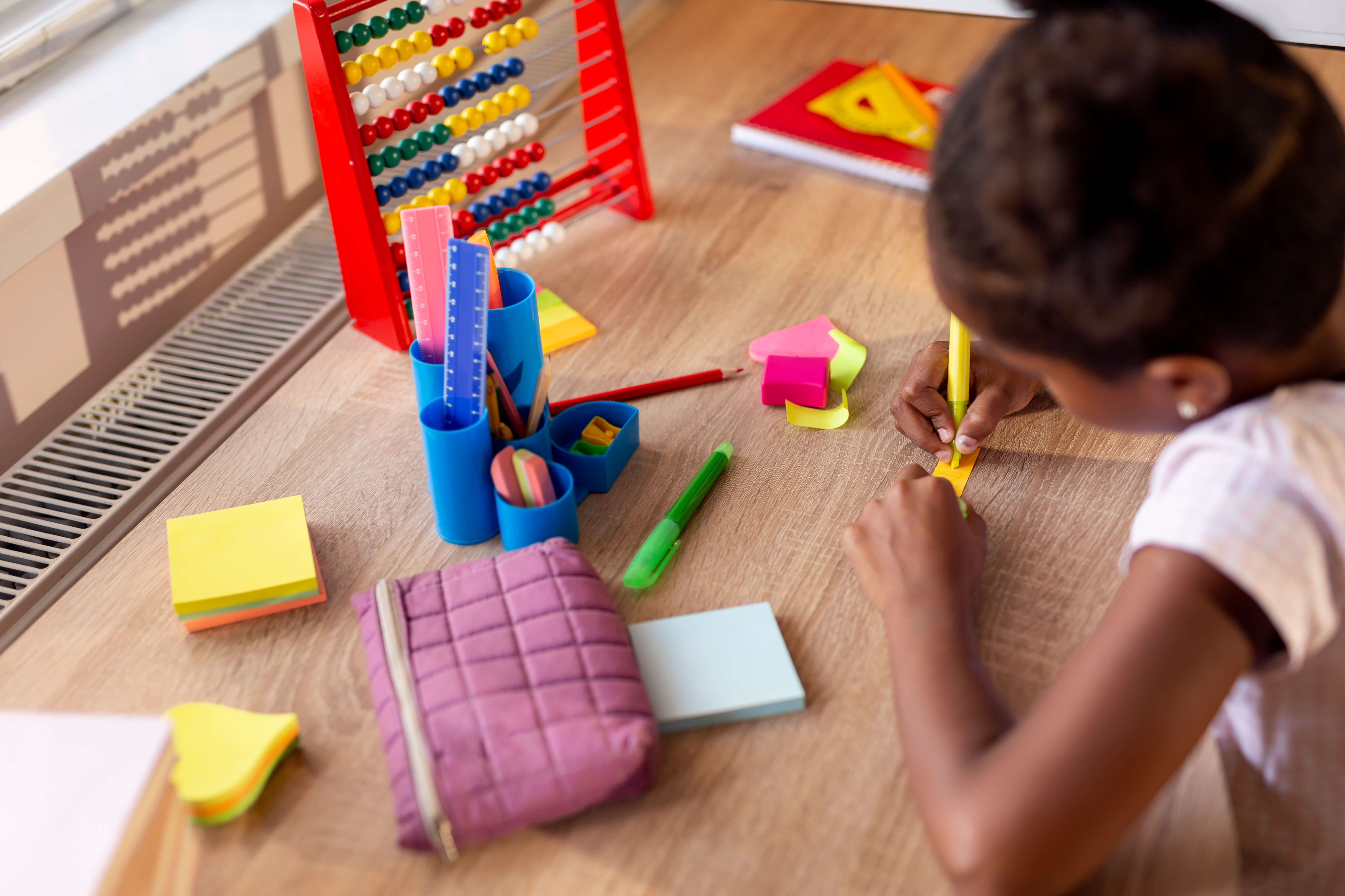 Primary-age girl writing on post-it notes and surrounded by maths equipment including a set square and abacus.